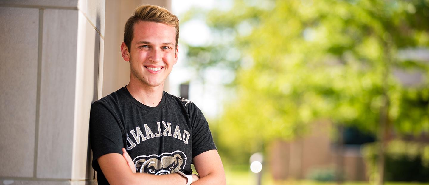 man in a black Oakland University t-shirt leaning on a wall outside