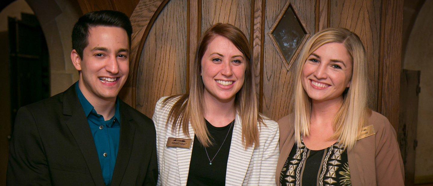 three smiling students, posing in front of a wooden door